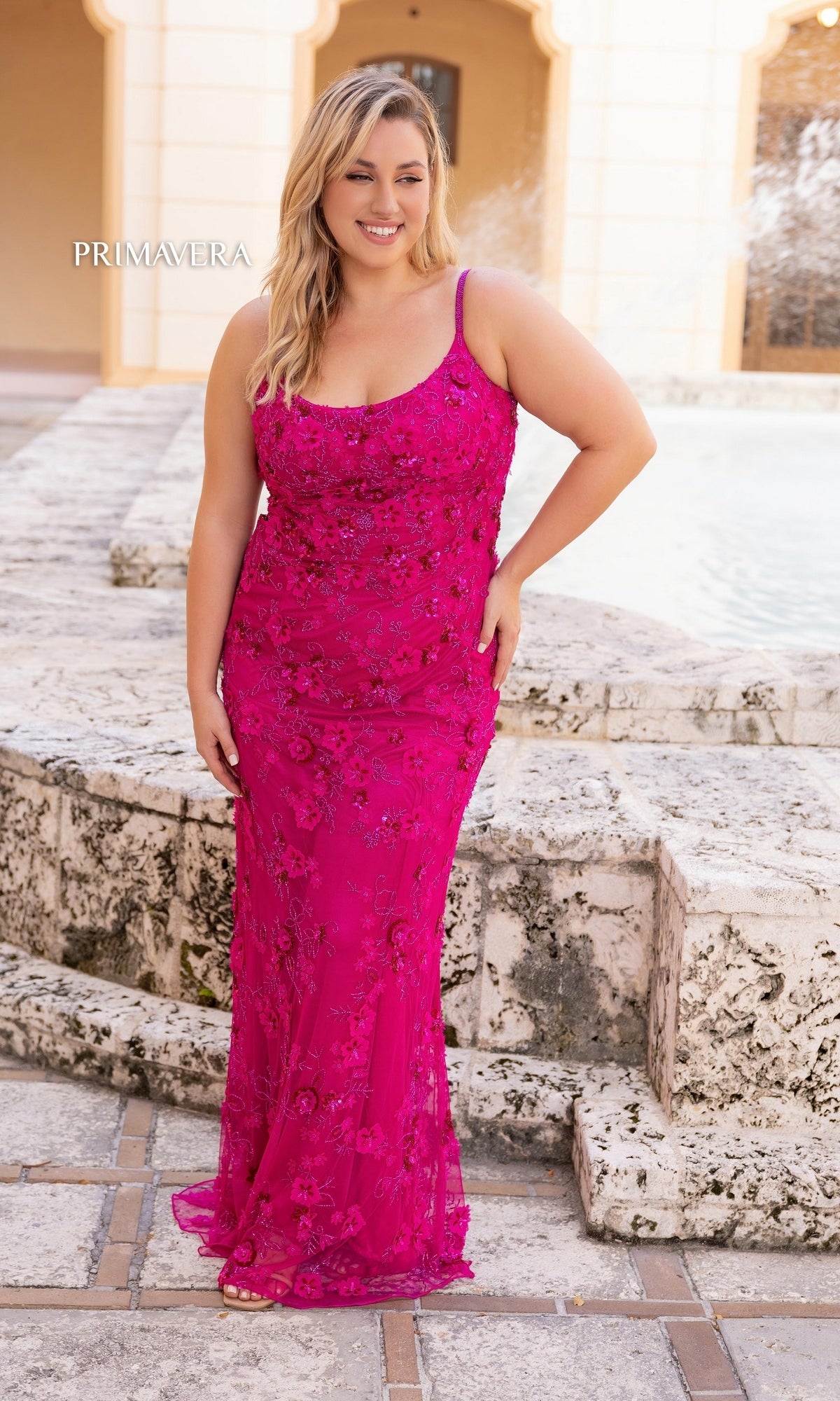 A woman wearing the Plus-Size Long Beaded Primavera Prom Dress 14050 in bright pink stands outdoors near stone steps and a fountain, smiling at the camera.